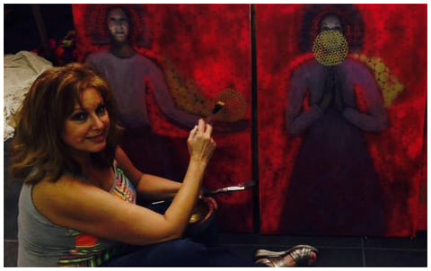 Auburn-haired woman in gray tank top sits on floor painting two large deep-red canvases of robed figures with hexagonal patterns