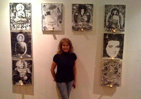 Woman in black shirt and jeans stands against cream gallery wall hung with sepia Buddha and portrait collage panels