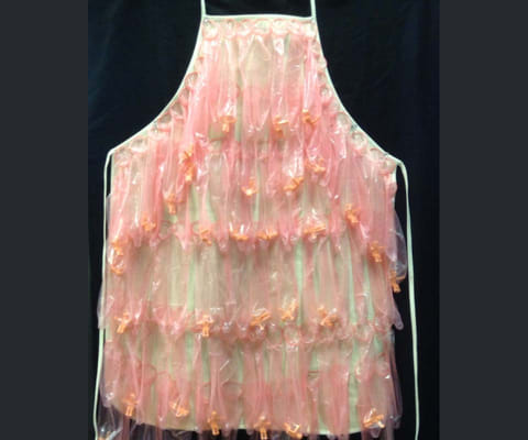 Translucent pink and clear plastic apron in ruffled tiers dotted with small orange satin bows, hung against a black backdrop