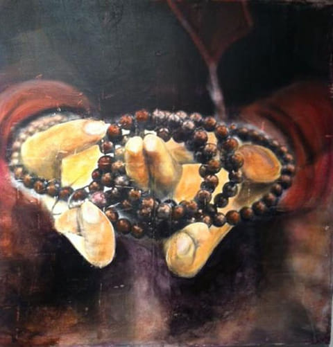 Two cupped hands painted in golden ochre holding a strand of dark brown prayer beads against a shadowy maroon background